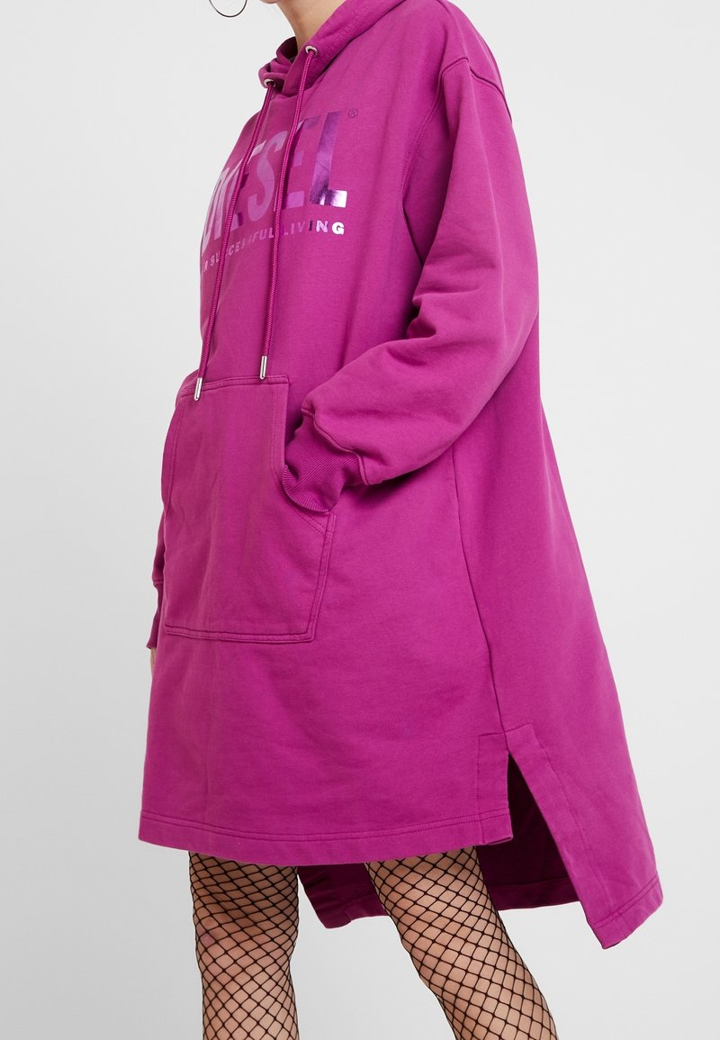 Person som bär en lysande lila oversized huvtröja med framficka och silvertext, tillsammans med svarta fiskenätsstrumpor mot en neutral bakgrund.