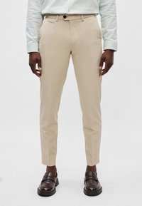 Μπεζ κομμένα παντελόνια με στενή εφαρμογή, μπροστινές τσέπες και φερμουάρ, σε συνδυασμό με σκούρες καφέ loafers, παρουσιασμένα σε άσπρο φόντο.