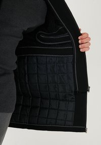 Giacca nera con interno trapuntato, dotata di cuciture argentate e una tasca con zip. Il materiale esterno appare morbido e strutturato.