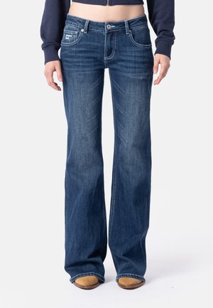 Person iført blå bootcut jeans med et lille frontlomme-mærke, kombineret med brune sko og en kort marineblå jakke.