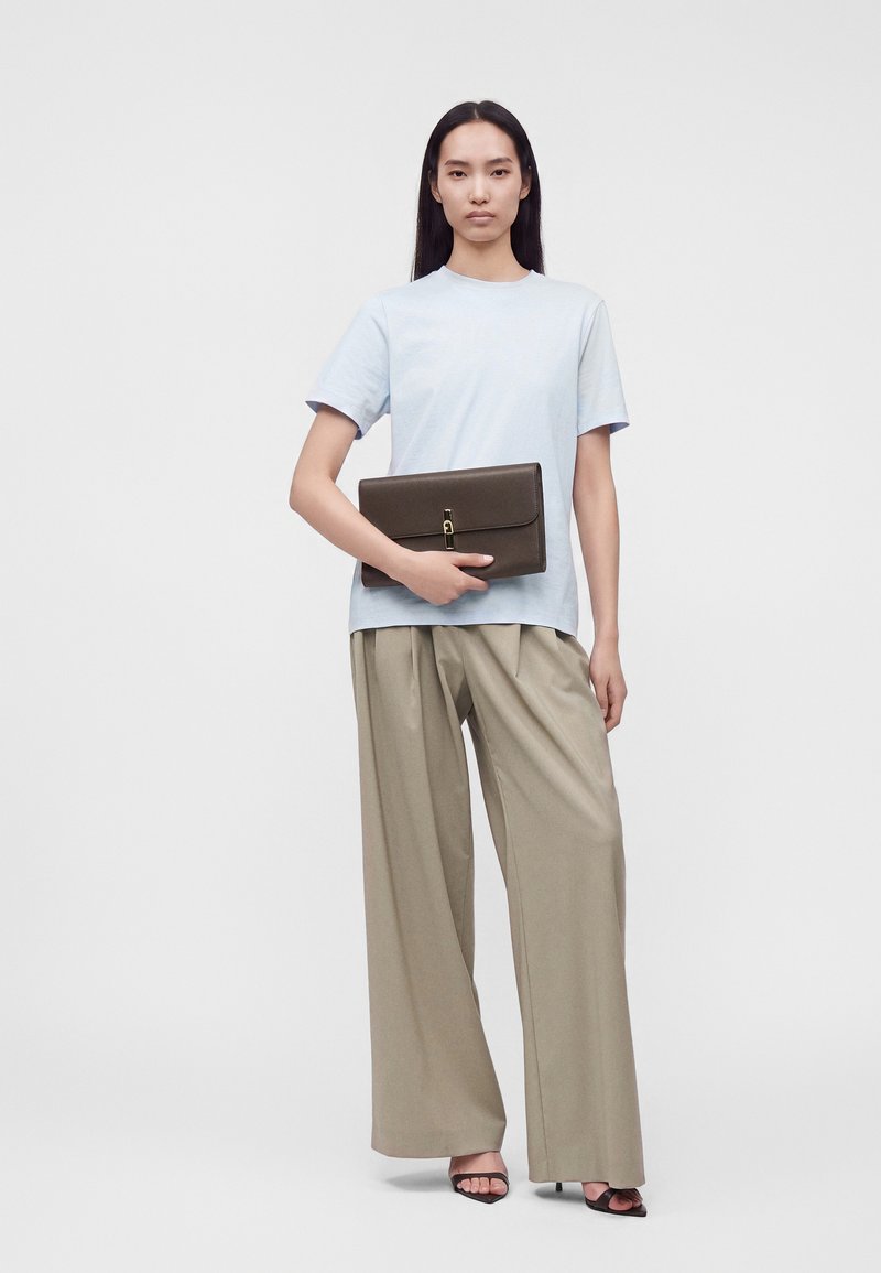 Femme portant un t-shirt bleu clair et un pantalon large beige, tenant une pochette marron foncé et portant des talons noirs ouverts.