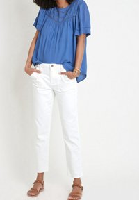 Modrá blůza s krátkým rukávem, s texturovaným vzorem a sklady, kombinovaná s bílými kalhotami rovného střihu a hnědými plochými sandály.