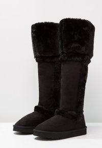 Svarta knähöga stövlar i konstpäls och mjukt mocka, med en fluffig krage, texturerade sidor och en platt gummisula.