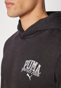 Haut à capuche noir avec une poche kangourou et un cordon de serrage, orné du logo "PUMA sports club" en blanc, graphique texturé sur la poitrine.