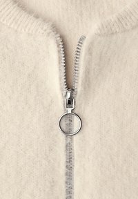 Jasnokremowy sweter zapinany na zamek błyskawiczny o puszystej fakturze, z metalicznym zamkiem z okrągłym uchwytem pośrodku przodu.