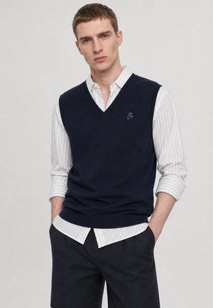 Ung mand med kort hår iført en navyblå sweatervest over en hvid stribet skjorte og mørke bukser med hænderne i lommerne.