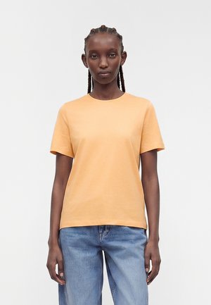 Jeune femme aux cheveux tressés porte un t-shirt uni pêche à manches courtes et un jean bleu clair, debout devant un fond blanc.