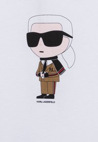 Karikatūra figūra brūnā uzvalkā ar melnu kaklasaiti un šalli, nēsājot lielas melnas saulesbrilles, izdrukāta uz baltas fona. "KARL LAGERFELD" teksts zemāk.