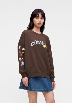 Femme aux longs cheveux foncés portant un sweat-shirt marron avec le texte "COMFY" et de petits graphismes, assorti à une jupe en jean bleue.