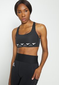 Sutiã desportivo preto com alças largas e padrões geométricos brancos na frente, combinado com leggings pretas de cintura alta. Textura suave.