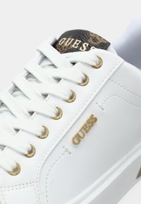 Baskets en cuir blanc avec lacets blancs, œillets dorés, logo "GUESS" doré sur le côté, et languette noire avec logos et texte dorés.
