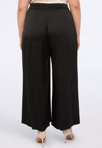 Černé široké kalhoty s elastickým pasem a hladkým, lesklým povrchem. Tkanina volně splývá až k kotníkům.