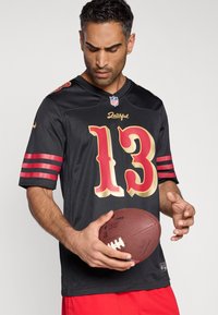 Μαύρη φανέλα NFL με κόκκινες και χρυσές λεπτομέρειες, με αριθμό 13 και τη λέξη «Faithful». Το μοντέλο κρατάει ένα ποδοσφαιρικό και φορά κόκκινο σορτς.