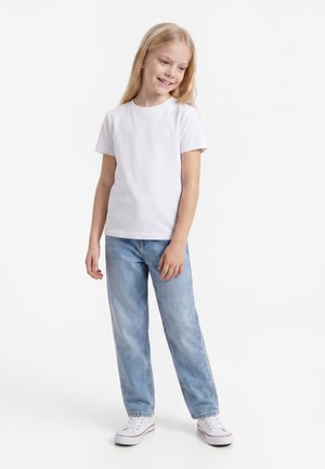 Jeune fille souriante aux longs cheveux blonds portant un t-shirt blanc uni, un jean bleu clair et des baskets blanches, debout devant un fond blanc.