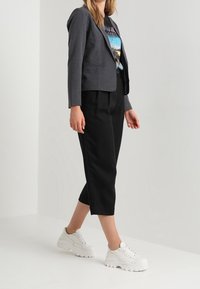 Blazer grigio su misura sopra una t-shirt grafica, abbinato a pantaloni neri a tre quarti e sneakers bianche. Dettagli visibili di bottoni e tasche.