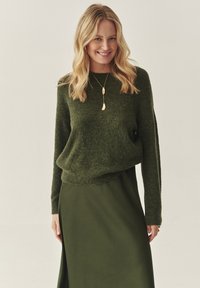 Zelený pletený svetr s kulatým výstřihem, s dlouhými rukávy a volným střihem, spárovaný s jednobarevnou tmavě zelenou sukní. Viditelný zlatý přívěsek na náhrdelníku.