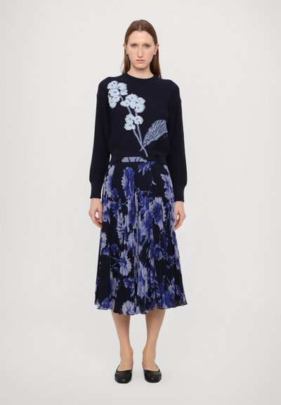 Navy knitted sweater with light blue floral embroidery paired with a pleated, floor-length skirt in navy and blue floral print, finished with flat shoes.