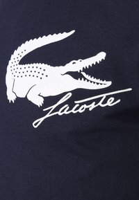 Námorní tkanina s bílým krokodýlím logem s detailními šupinami a kurzívou "Lacoste" pod ním. Hladký povrch, bez vzorů.