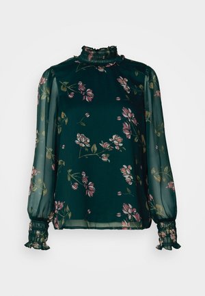 Blouse florale verte à manches longues, présentant un col haut à volants et des poignets élastiqués, fabriquée en tissu léger et transparent avec des fleurs roses et beiges.