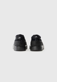 Pair of black UGG shoes viewed from the back, featuring pull tabs with toggle closures and thick rubber soles on a plain white background.