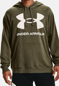 Homme portant un sweat à capuche Under Armour vert olive avec un grand logo blanc et une poche avant, associé à un pantalon noir avec des détails blancs.