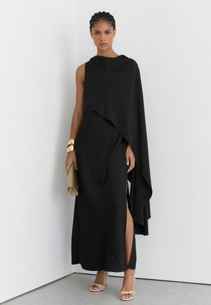 Frau mit geflochtenem Haar, die ein ärmelloses schwarzes Kleid mit einem drapierten Umhang und seitlichem Schlitz trägt, hält eine goldene Clutch und trägt goldene Armbänder.
