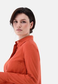 Long-sleeve, collared shirt in a textured rust color, featuring a button placket and a chest pocket. Smooth fabric, fitted silhouette.