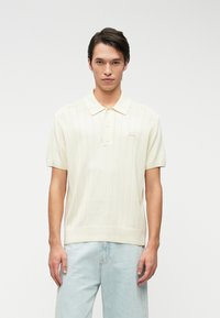 Κοντομάνικο polo σε κρέμα απόχρωση με κάθετη ραφή, κλείσιμο με δύο κουμπιά και κεντητό λογότυπο στο στήθος. Συνδυασμένο με ανοιχτό μπλέ τζιν.