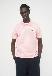 Ανοιχτό ροζ πόλο μπλούζα με λευκές ρίγες, κοντά μανίκια, γιακά και ένα μαύρο και κίτρινο λογότυπο κεντημένο στο στήθος.