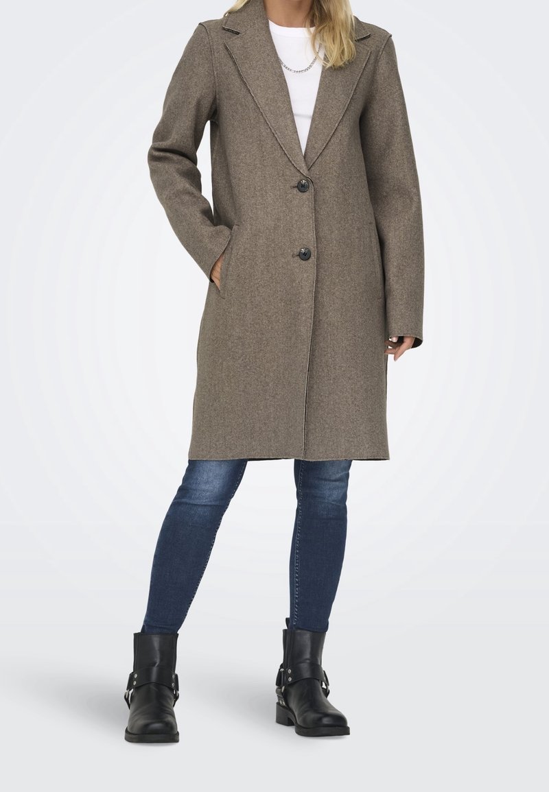 Manteau marron en mélange de laine avec un col cranté, deux poches avant et deux boutons noirs. Assorti à un jean bleu et des bottines noires.