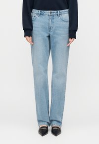 Šviesiai mėlynos spalvos džinsai iš džinso audinio, tiesaus kirpimo, su penkiomis kišenėmis ir vidutinio aukščio liemeniu. Dėvimi su juodais bateliais su aštriomis noselėmis.