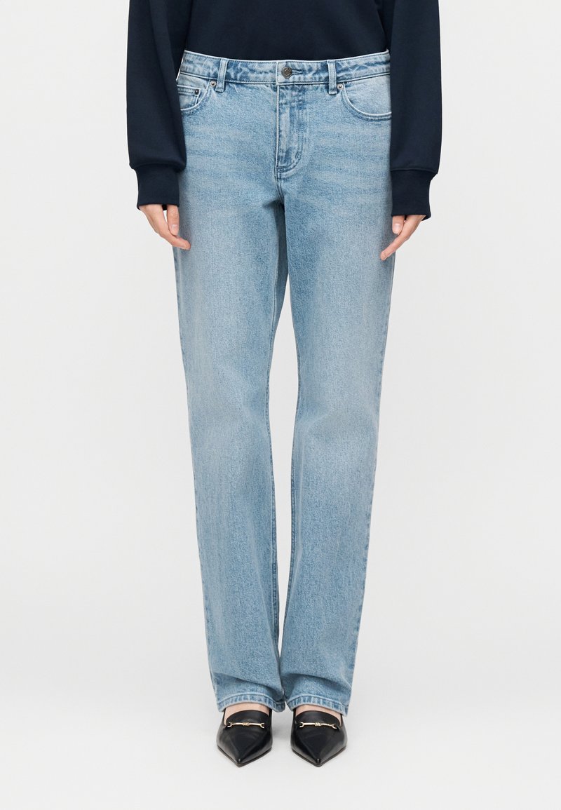 Šviesiai mėlynos spalvos džinsai iš džinso audinio, tiesaus kirpimo, su penkiomis kišenėmis ir vidutinio aukščio liemeniu. Dėvimi su juodais bateliais su aštriomis noselėmis.