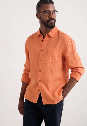 Orange linen button-up shirt with a chest pocket, long sleeves rolled to the elbows, and brown buttons, paired with dark trousers.