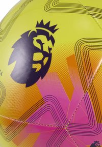 Ronde voetbal met een heldergele oppervlakte, zwart leeuwlogo en levendige roze en oranje geometrische patronen met gebogen lijnen.