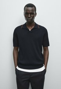 Czarny krótki rękaw polo sweter o pionowym żebrowanym wzorze i brązowym akcentem na kołnierzyku, noszony na białej koszuli, w połączeniu z ciemnymi spodniami.