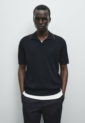 Sort kortærmet polo sweater med vertikal ribstrik og en brun detalje ved kraven, lagd over en hvid skjorte, båret med mørke bukser.