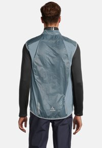 Leichte blaue Weste aus glattem Material, mit hohem Kragen, schwarzen langen Ärmeln darunter und reflektierenden Akzenten auf dem Rücken.