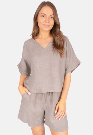 Femme aux longs cheveux portant un haut ample taupe à col en V et un short assorti, debout avec une main dans la poche, fond clair.