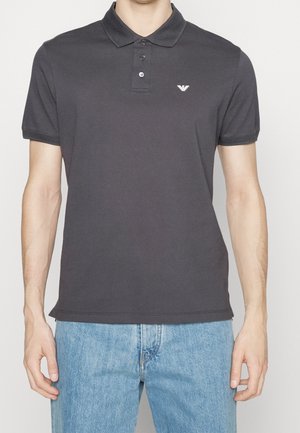Mann trägt ein dunkelgraues kurzärmeliges Polo mit kleinem weißem Logo auf der linken Brustseite und hellblaue Jeans.