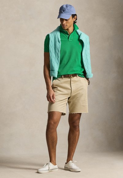 Άνδρας με πράσινο πόλο πουκάμισο, μπεζ σορτς, ανοιχτό μπλε πουλόβερ περασμένο στους ώμους, μπλε καπέλο και λευκά αθλητικά παπούτσια, στέκεται μπροστά σε ουδέτερο φόντο.