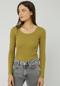 Haut à manches longues, de couleur vert olive, avec un col arrondi, accompagné d'un jean taille haute délavé gris clair avec une fermeture à boutons.