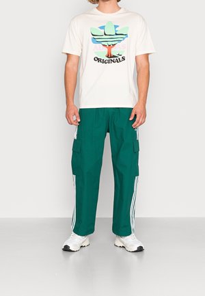 Mężczyzna ubrany w białą koszulkę z grafiką Adidas Originals, zielone spodnie dresowe Adidas z białymi paskami i białe sneakersy, stojący na szarym podłożu.