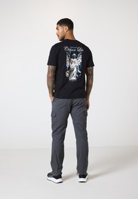 Schwarzes Baumwoll-T-Shirt mit einem detaillierten Grafikum eines geflügelten Körpers auf dem Rücken. Getragen mit grauen Cargo-Hosen und weißen Sneakers.