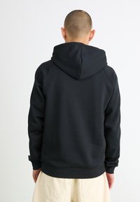 Pessoa com a cabeça rapada, vestindo um hoodie preto e calças bege, de costas contra um fundo branco simples.