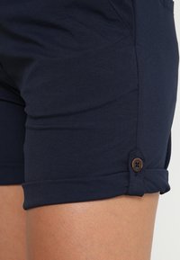 Tmavě modré bavlněné šortky se zaobleným lemem a hnědým knoflíkovým detailem. Tkanina má hladkou strukturu a jednoduchý jednobarevný design.