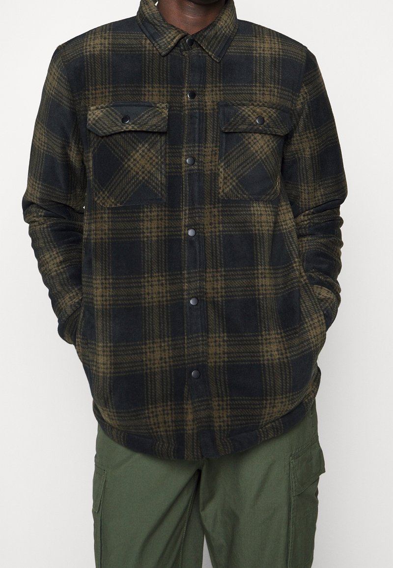 Homme portant une chemise à carreaux noir et vert olive à boutons avec poches à rabat sur la poitrine et pantalon cargo vert, les mains dans les poches.