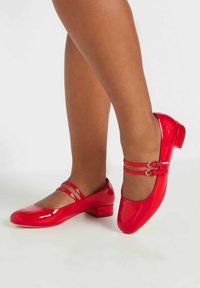 Punaiset lakatut ballerinat, joissa on pyöristetty kärki ja kaksi koristeellista hihnaa, alhaisen lohko-ireesein ja sileä, kiiltävä pinta.