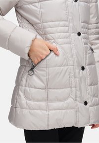 Jasnoszara kurtka puchowa z pikowanym wzorem, z czarnymi zapięciami na guziki i zamykana kieszenią boczną. Gładki, izolowany materiał.