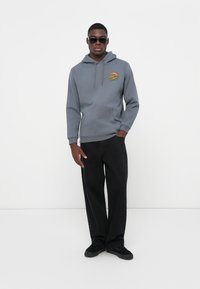 Felpa grigia con cappuccio e tasca frontale, caratterizzata da una grafica colorata. Abbinate a pantaloni larghi neri e scarpe da ginnastica scure.