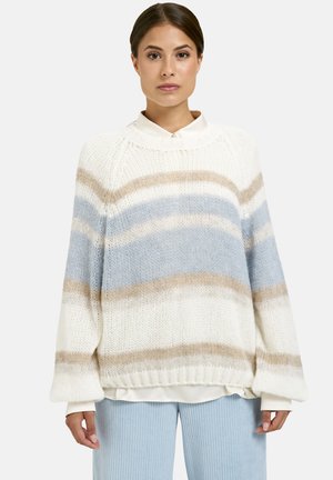 Eine Frau trägt einen weißen Pullover mit beigen und hellblauen horizontalen Streifen, der über einem weißen Hemd liegt, kombiniert mit hellblauen gerippten Hosen.